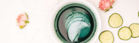 top view of bowl with lotion and cotton pads near tea roses and cucumber slices on white, bannerの写真素材
