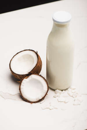 bottle of coconut milk near halves and flakes on white surface isolated on black