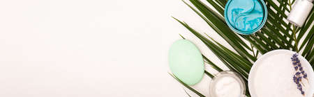 top view of bottle with coconut milk, soap, containers with cosmetic cream and scrub near palm...