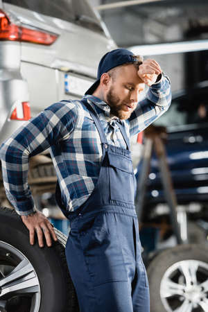 exhausted mechanic holding hand near forehead while standing near car wheel with closed eyes on blurred backgroundの写真素材