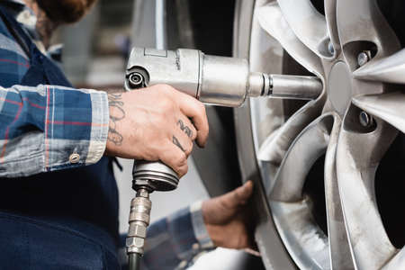 cropped view of mechanic fixing car wheel with pneumatic wrench on blurred background