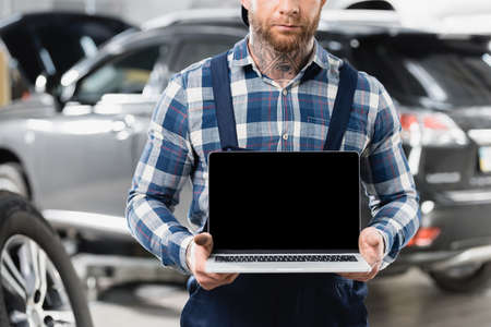 cropped view of technician holding laptop with blank screen near automobile on blurred background