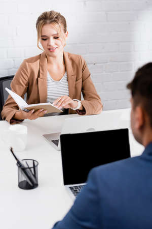 Smiling businesswoman looking at notebook, while sitting at table with laptops in office on blurred foregroundの写真素材