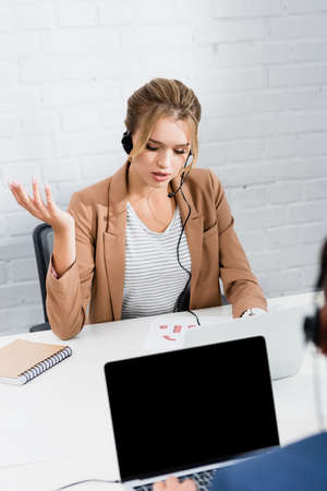 Female consultant in headset gesturing, while sitting at table in office on blurred foreground