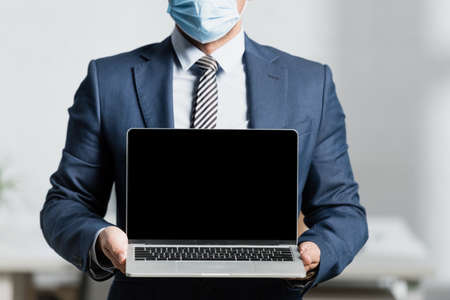 Cropped view of executive in medical mask, showing laptop with blank screen on blurred backgroundの写真素材