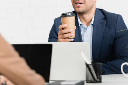 Cropped view of office worker with paper cup talking, while sitting at table with laptops on blurred foregroundの写真素材