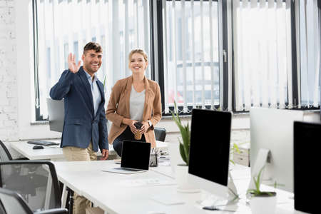 Happy businessman with waving hand, looking at camera, while standing near co-worker during break in officeの写真素材