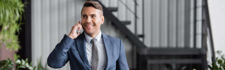 Smiling businessman looking away, while talking on mobile phone at work on blurred background,...