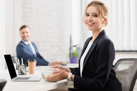 Smiling executive with meal in plastic bowl looking at camera, while sitting near laptop at table on...