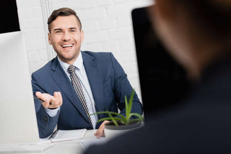 Happy executive gesturing, while sitting at workplace with blurred female co-worker on foregroundの写真素材