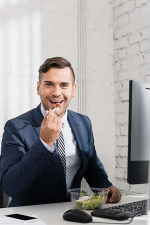 Smiling businessman looking at camera, while eating meal from plastic bowl at workplaceの写真素材