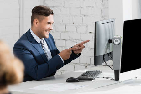 Cheerful businessman pointing with fingers while looking at laptop at workplace on blurred foregroundの写真素材