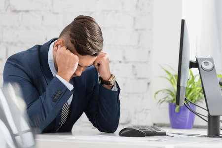 Tired businessman with hands near head, sitting near computer at workplace on blurred foreground