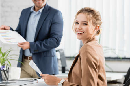 Happy businesswoman with document looking at camera, while sitting at workplace with blurred colleague on backgroundの写真素材