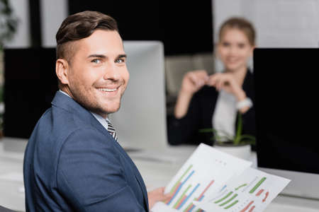 Smiling businessman with paper sheets looking at camera, while sitting at workplace with blurred co-worker on backgroundの写真素材
