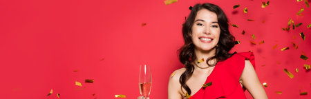 Smiling woman holding glass of champagne under falling confetti on red background, bannerの写真素材