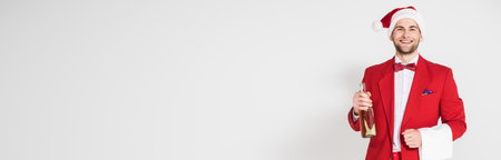 Smiling man in suit and santa hat holding towel and bottle of champagne on gray background, bannerの写真素材