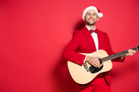 Smiling man in santa hat playing acoustic guitar on red backgroundの写真素材