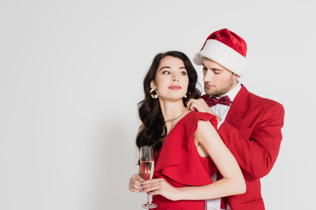 Man in santa hat and bow tie wearing necklace on woman in dress with glass of champagne on gray backgroundの写真素材