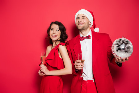 Smiling couple with glasses of champagne, disco ball and santa hat on red backgroundの写真素材