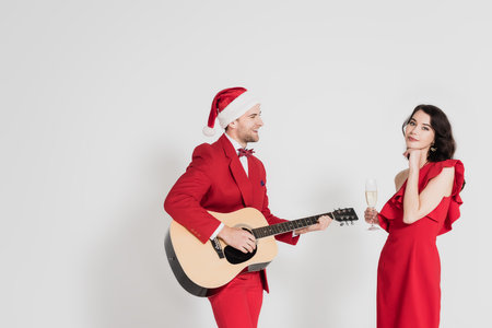 Smiling man in sata hat playing acoustic guitar near woman with glass of champagne on gray backgroundの写真素材