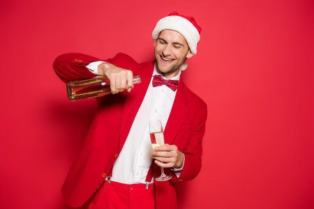 Smiling man in santa hat and suit pouring champagne in glass on red backgroundの写真素材