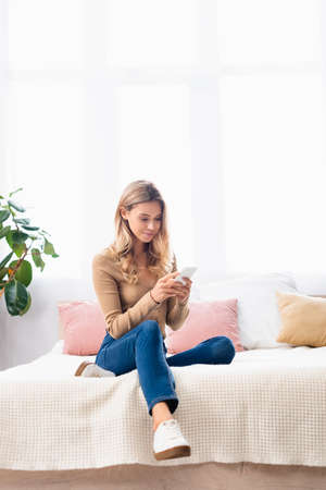 Young woman in casual clothes using smartphone on bed