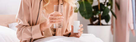 Cropped view of woman holding glass of water and jar with pills on bed, banner