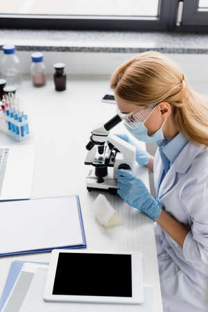 scientist in medical mask and goggles looking through microscope on desk