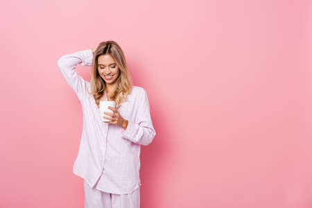 Smiling woman in pajamas with hand near head holding cup on pink backgroundの写真素材