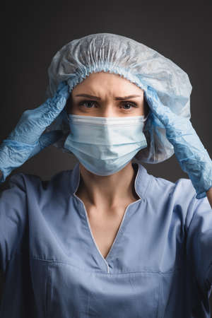 nurse in medical cap and mask suffering from headache isolated on dark gray
