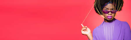 african american young woman in purple stylish outfit and sunglasses with yellow strings on face isolated on red, bannerの写真素材