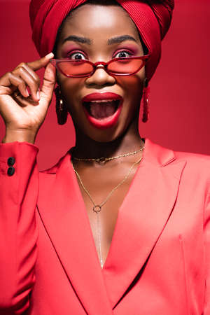 excited african american young woman in stylish outfit, sunglasses and turban isolated on red