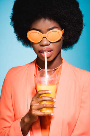 african american young woman in stylish outfit with orange juice isolated on blue backgroundの写真素材