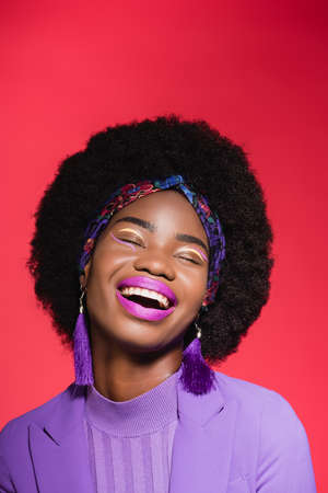 laughing african american young woman in purple stylish outfit isolated on red