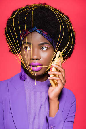 african american young woman in purple stylish outfit with yellow strings on face holding smartphone...
