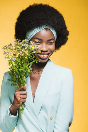 smiling african american young woman in blue stylish outfit with chamomile bouquet isolated on yellowの写真素材