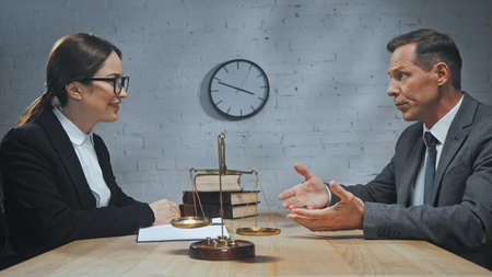 Smiling insurance agent looking at colleague pointing with hands near notebook, scales and books on...
