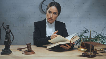 Insurance agent looking at camera while holding book near statuette of justice, gavel and scales