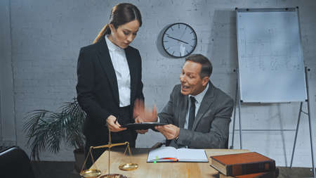 Aggressive insurance agent holding clipboard near colleague, notebook and scales in officeの写真素材