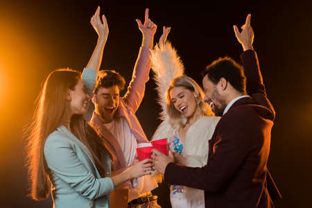 group of excited multicultural friends toasting plastic cups and rejoicing during party on blackの写真素材