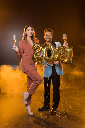 full length of happy multicultural couple holding glasses of champagne near balloons with 2021...