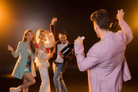 back view of man holding bottles with champagne near excited multicultural friends on black