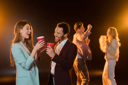 cheerful woman and african american man holding plastic cups near friends dancing during party on...