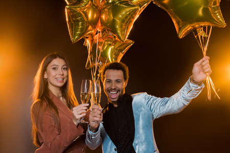smiling interracial couple toasting glasses with champagne and holding balloons on black