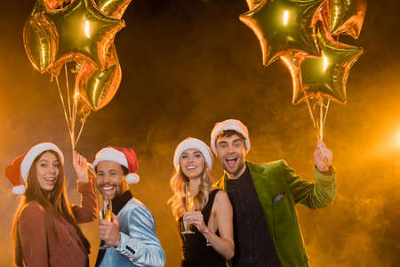 smiling multicultural friends in santa hats holding glasses with champagne near balloons on black