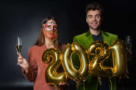 happy man holding balloons with 2021 numbers near woman in carnival mask with glass of champagne on blackの写真素材