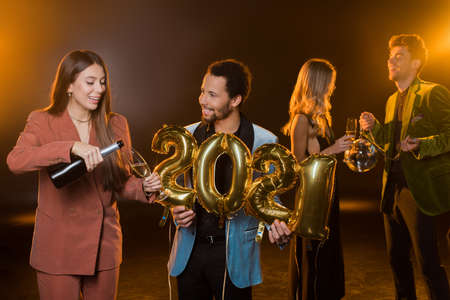happy woman pouring champagne in glass near african american man and friends on blurred and black backgroundの写真素材