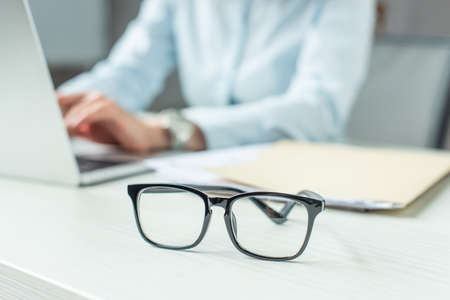 Cropped view of businesswoman typing on laptop, while sitting at table with eyeglasses on blurred backgroundの写真素材