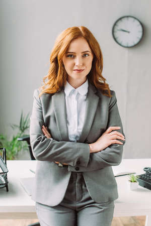 Front view of positive businesswoman with crossed arms looking at camera, while leaning on table on blurred backgroundの写真素材